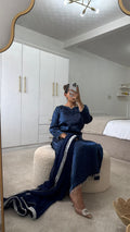 Woman in a blue long qameez with an embellished dupatta seated on a cream ottoman in a stylish room.