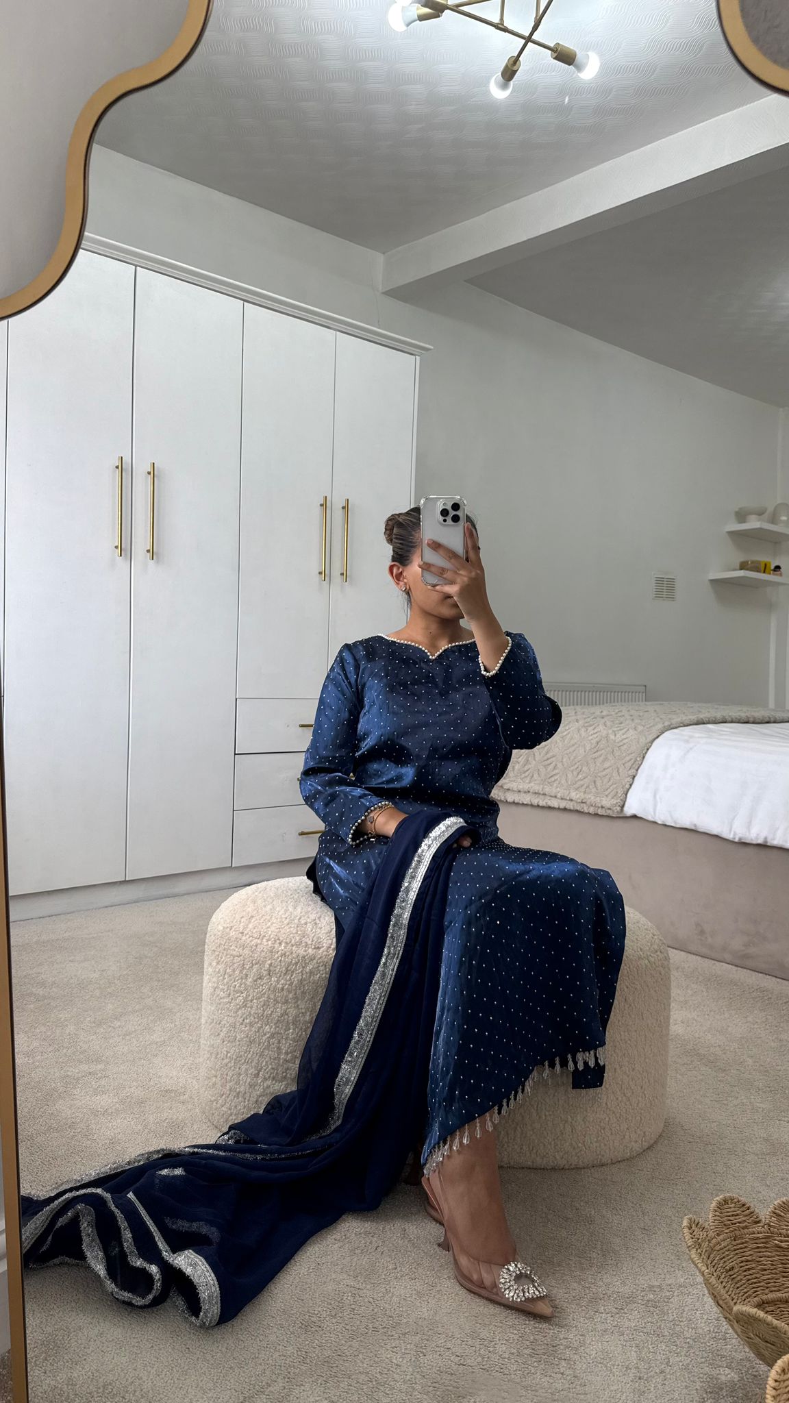 Woman in a blue long qameez with an embellished dupatta seated on a cream ottoman in a stylish room.
